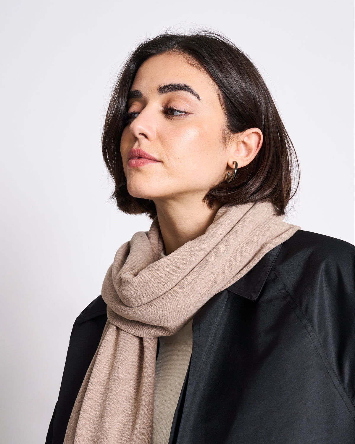 A woman with short dark hair wears the jan-n-june SCARF ROSS SAND, a black coat, and gold hoop earrings. She looks to the side with a calm expression against a plain white background.