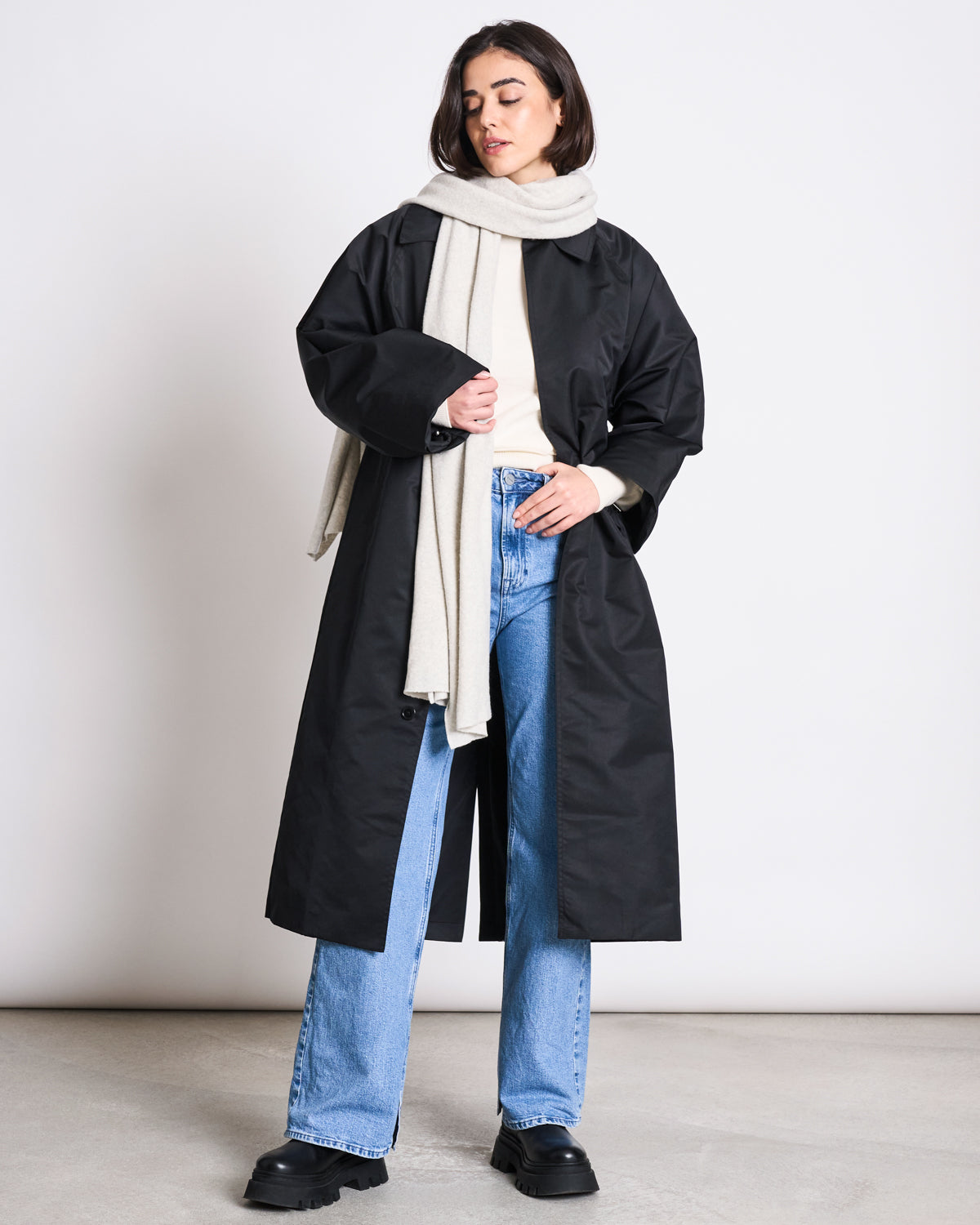 A woman stands against a plain background wearing a long black coat, light blue jeans, black shoes, a white turtleneck, and the jan-n-june SCARF ROSS LIGHT GREY vegan scarf draped around her neck.