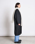 A woman stands in profile against a plain background, wearing a long black coat, blue jeans, black platform shoes, and the jan-n-june SCARF ROSS LIGHT GREY draped cozy around her neck.