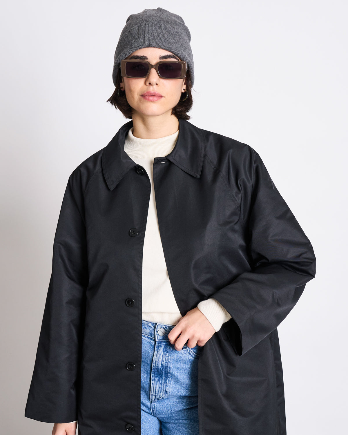 A person wearing the jan-n-june KNIT BEANIE BROOKLYN DARK GREY GOTS, dark sunglasses, a black coat, a white sweater, and blue jeans stands against a plain light background, looking confidently at the camera.