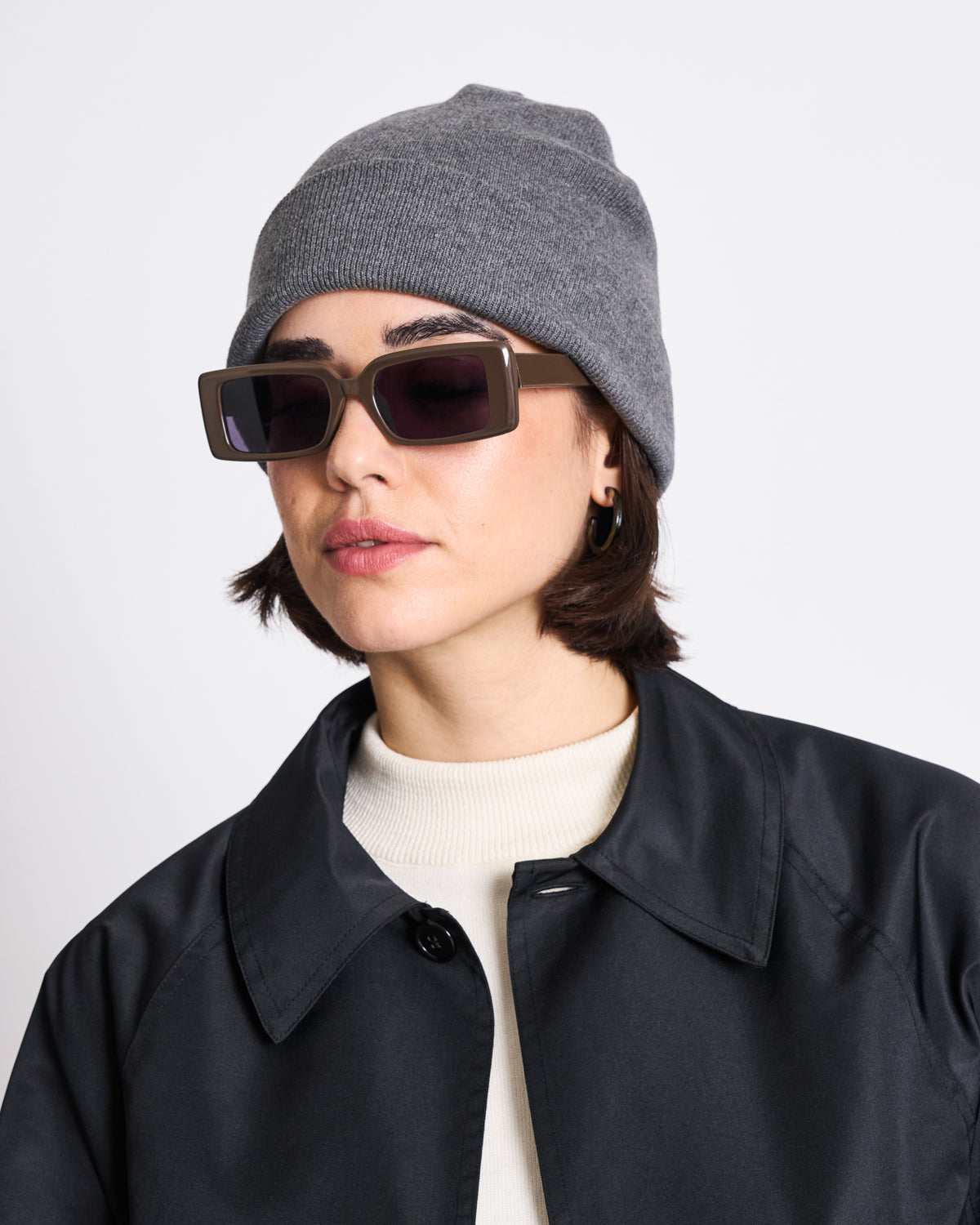 A woman wears the jan-n-june KNIT BEANIE BROOKLYN DARK GREY GOTS with sunglasses, a black jacket, and a white top, looking slightly to the side against a plain white background.
