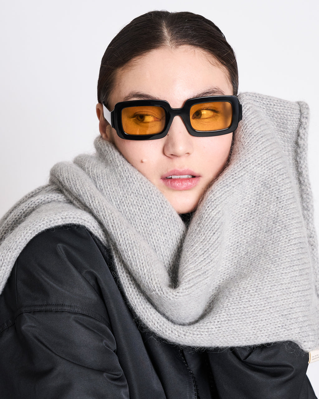 A woman wearing rectangular amber-tinted sunglasses, a black jacket, and the jan-n-june WOOLY KNIT SCARF AVAK LIGHT GREY looks to the side against a plain light background.