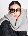 A woman wearing rectangular amber-tinted sunglasses, a black jacket, and the jan-n-june WOOLY KNIT SCARF AVAK LIGHT GREY looks to the side against a plain light background.