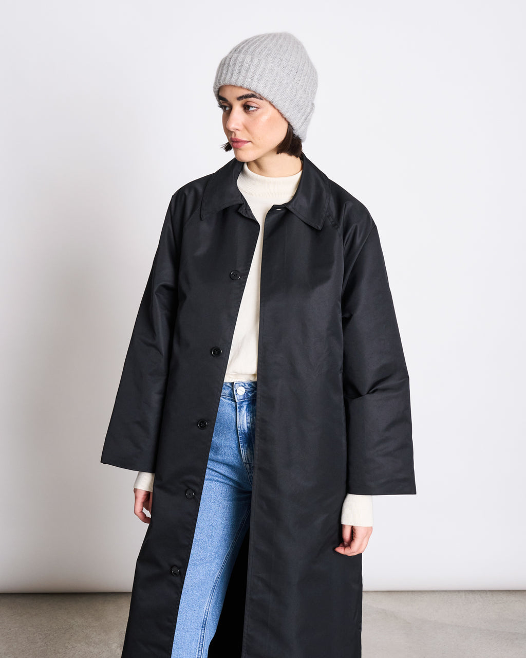 A woman in a long black coat, light blue jeans, a white sweater, and the jan-n-june WOOLY KNIT BEANIE BJON LIGHT GREY stands against a plain white background, gazing slightly to her left.