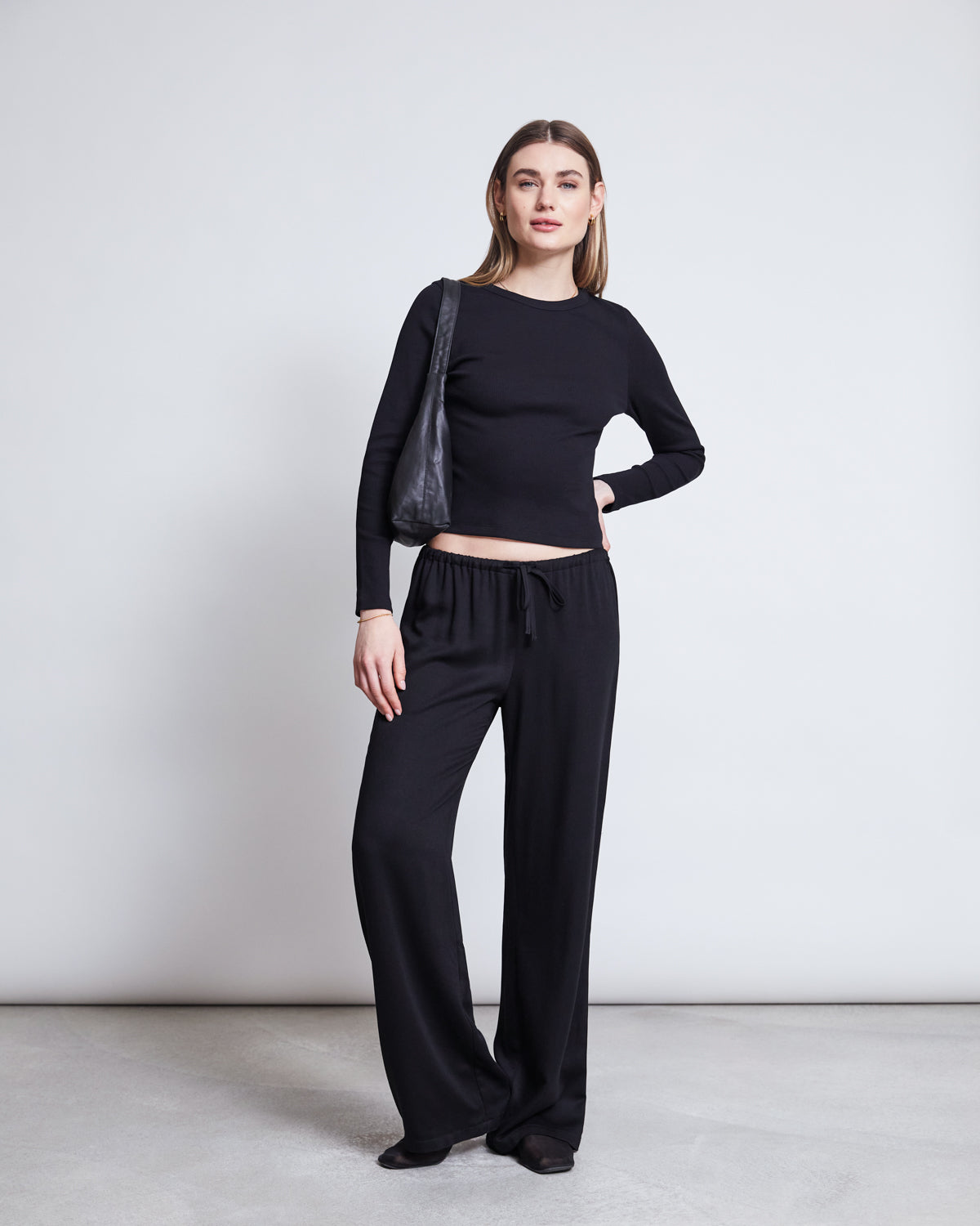 A woman poses confidently against a light background wearing jan-n-junes MID RISE LOOSE PANTS STRATH BLACK and a long-sleeve black MicroModal top, accessorized with a black shoulder bag.