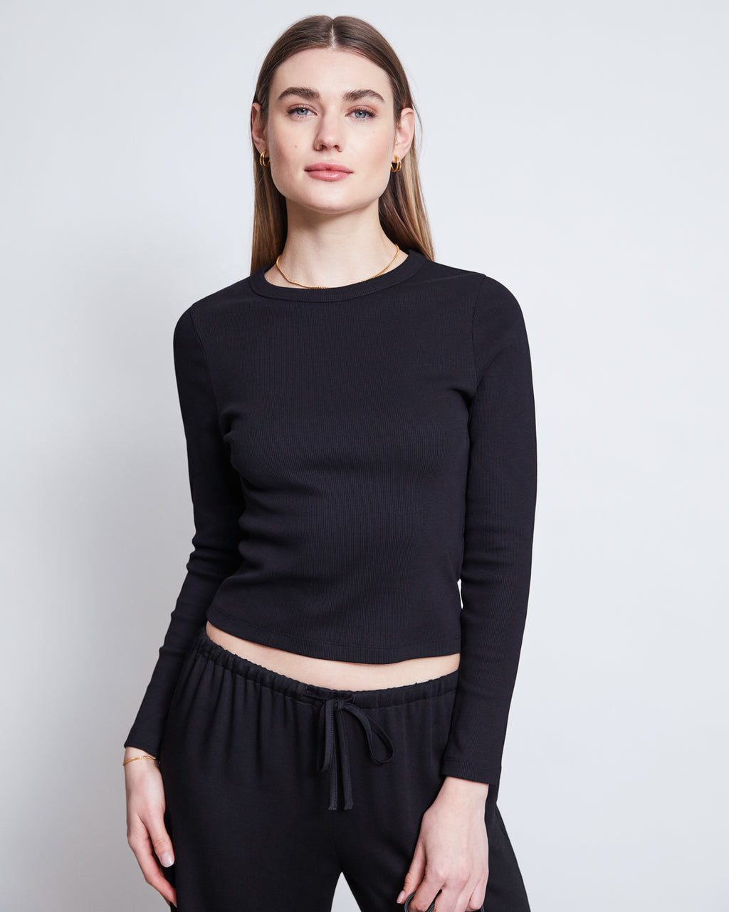 A woman with straight, light brown hair wears jan-n-june’s MID RISE LOOSE PANTS STRATH BLACK and a fitted black long-sleeve top, standing against a plain light background and looking at the camera with a neutral expression.