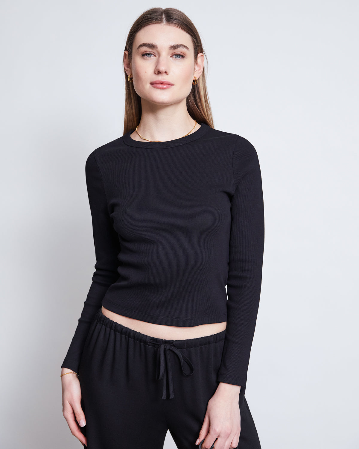 A woman with straight, light brown hair wears jan-n-june’s MID RISE LOOSE PANTS STRATH BLACK and a fitted black long-sleeve top, standing against a plain light background and looking at the camera with a neutral expression.