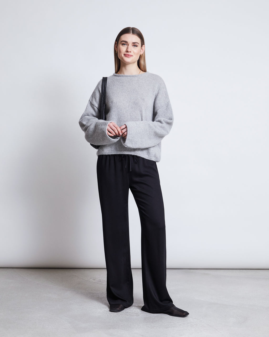A person stands against a plain light background, smiling gently. They wear jan-n-june’s MID RISE LOOSE PANTS STRATH BLACK—crafted from soft EcoVero fabric—with a loose gray sweater, black shoes, a black shoulder bag, and straight brown hair.