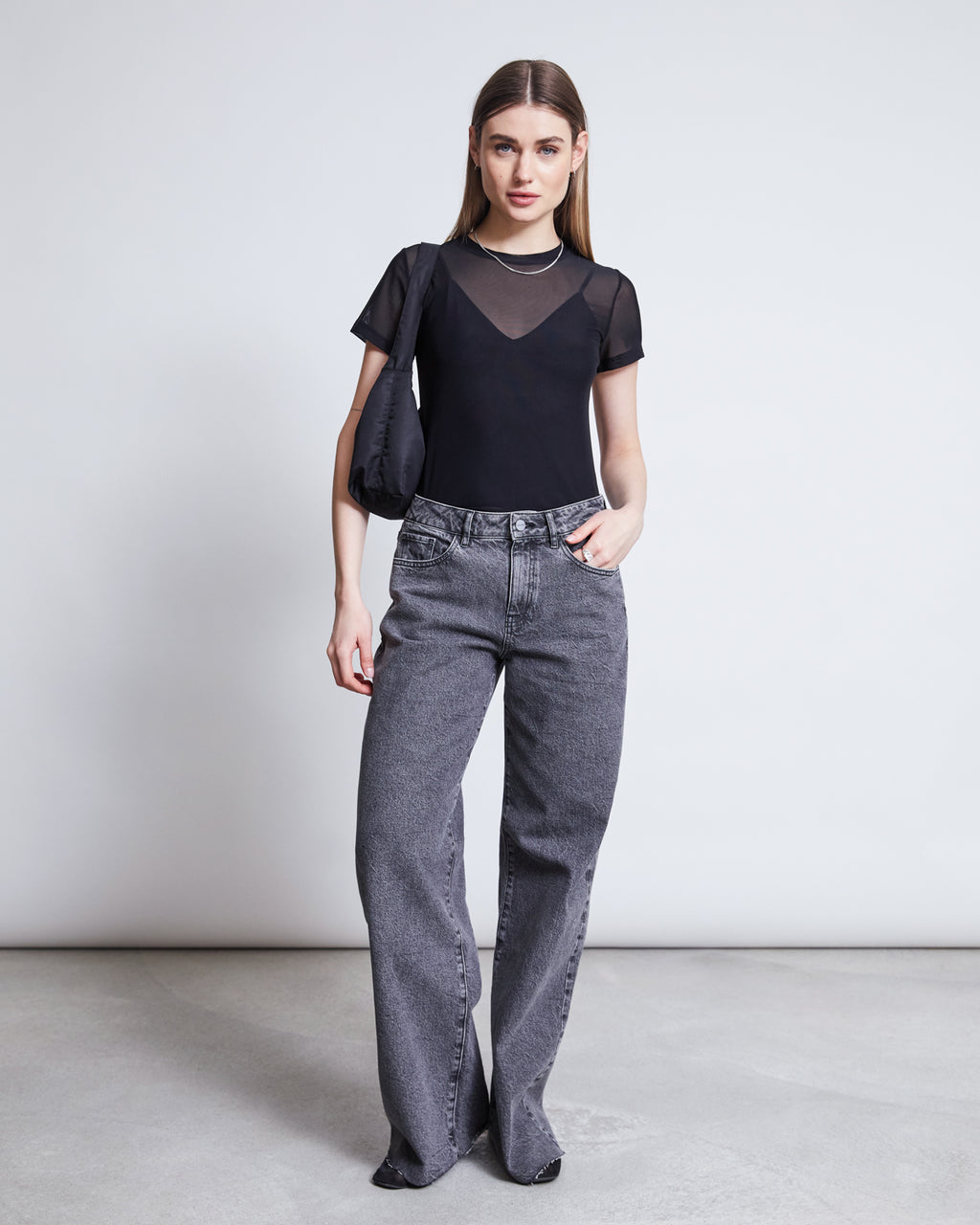 A woman with straight brown hair and a neutral expression wears the jan-n-june MESH T-SHIRT EVIE BLACK over a black camisole, paired with high-waisted loose gray jeans and a black shoulder bag—an ideal festival outfit against a plain background.