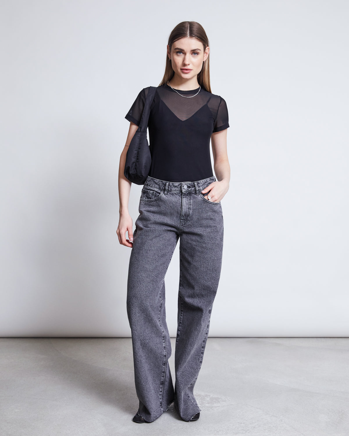 A woman with straight brown hair and a neutral expression wears the jan-n-june MESH T-SHIRT EVIE BLACK over a black camisole, paired with high-waisted loose gray jeans and a black shoulder bag—an ideal festival outfit against a plain background.