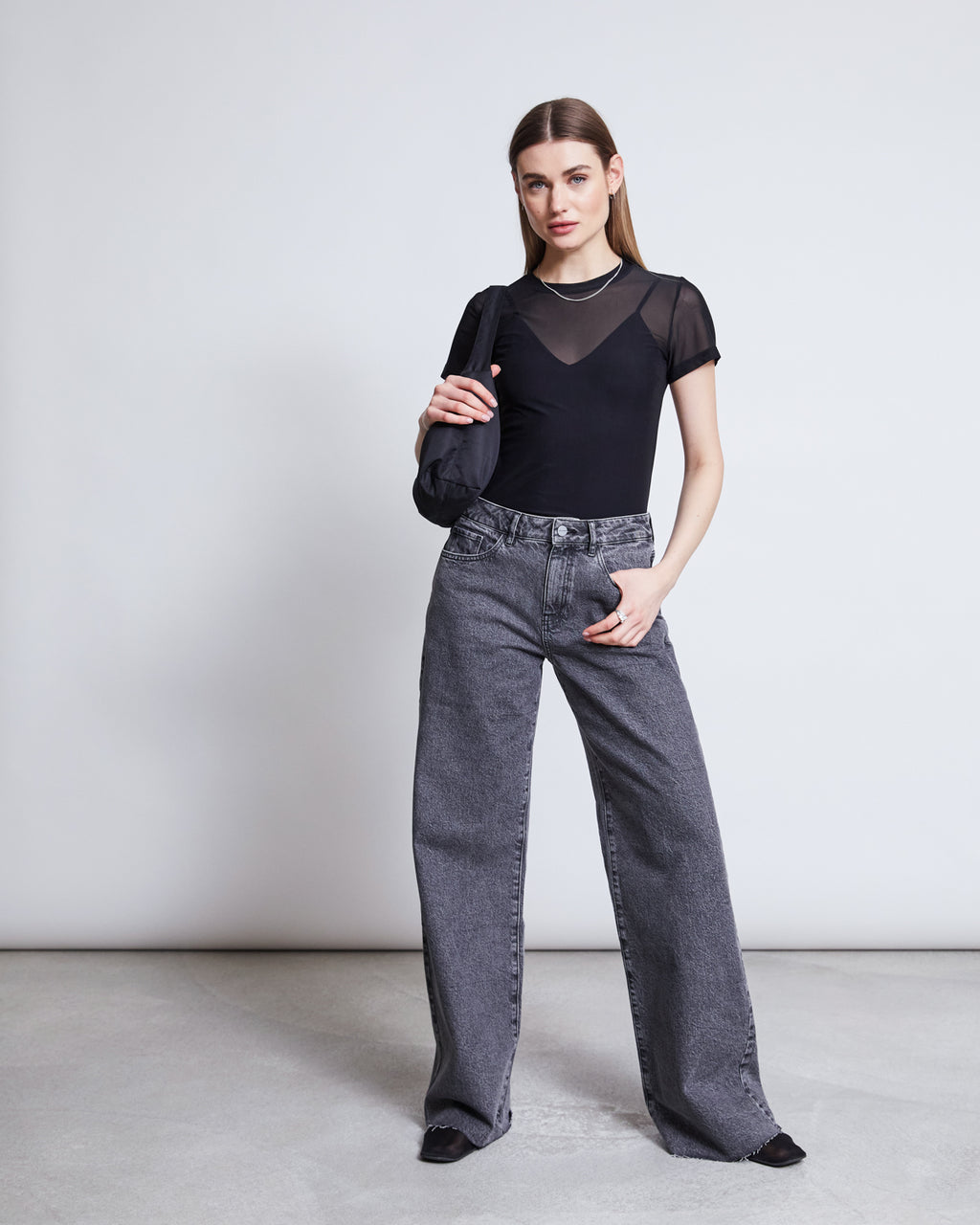 A woman stands against a plain background wearing the jan-n-june MESH T-SHIRT EVIE BLACK, paired with gray wide-leg jeans, black shoes, and a small black shoulder bag—ideal for festivals or casual outings.