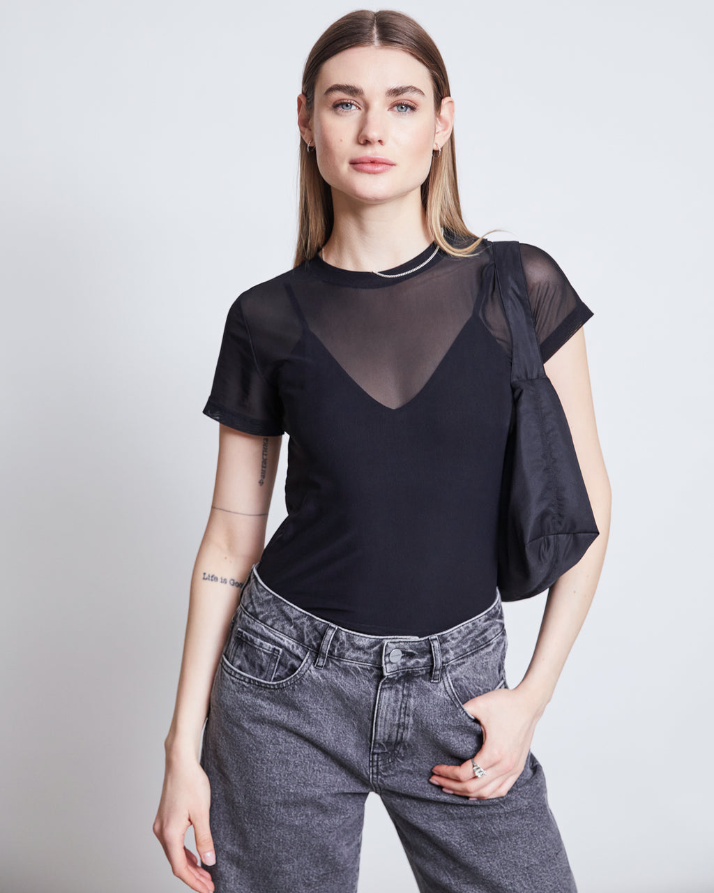 A woman with long straight hair wears the jan-n-june MESH T-SHIRT EVIE BLACK over a black bralette, gray jeans, and carries a black shoulder bag, posing with one hand in her pocket—ideal for festivals or casual outings.