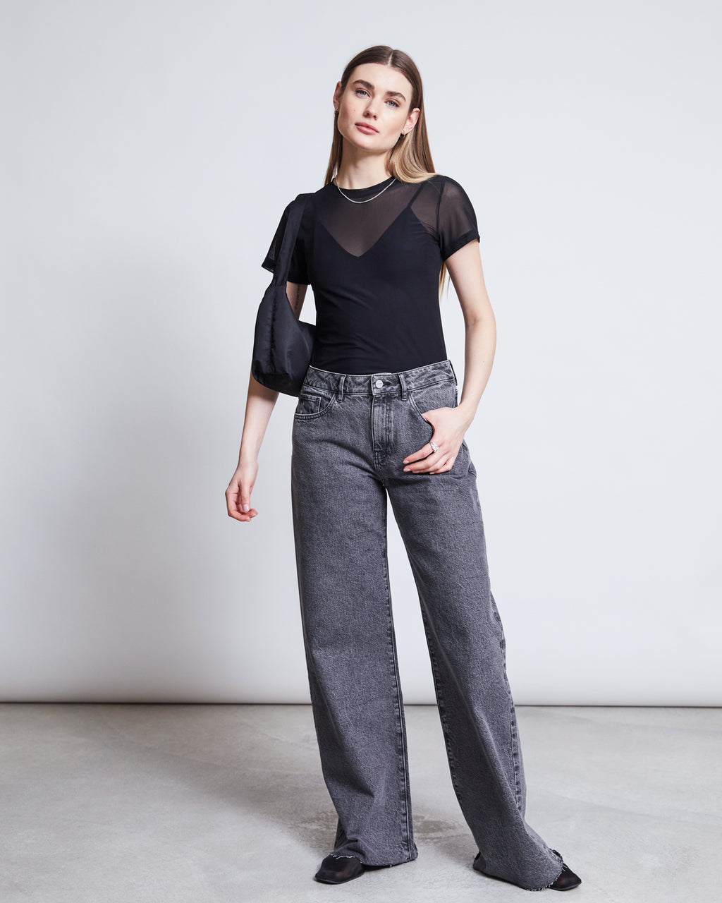 A woman with straight hair and a neutral expression wears the jan-n-june MESH T-SHIRT EVIE BLACK over a tank, paired with loose gray jeans, black shoes, and a black shoulder bag—perfect for festival style.