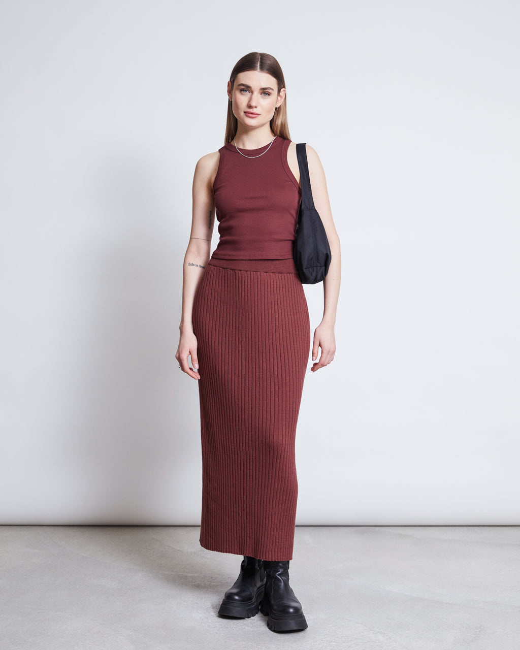 A woman stands against a plain light background, wearing the jan-n-june MIDI KNIT SKIRT EBBA INTENSE RUST GOTS with a sleeveless maroon top. She has straight hair, black boots, and carries a black shoulder bag.