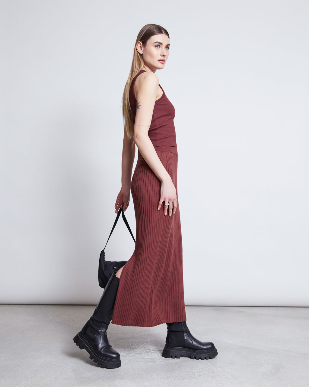 A woman with long, straight hair wears jan-n-june’s MIDI KNIT SKIRT EBBA in intense rust, styled with black chunky boots and a small black handbag, standing against a plain light gray background.