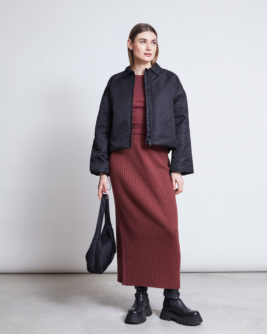 A woman with straight brown hair and a neutral expression stands against a plain background wearing the jan-n-june MIDI KNIT SKIRT EBBA INTENSE RUST GOTS, paired with a black jacket, chunky boots, and holding a black handbag.