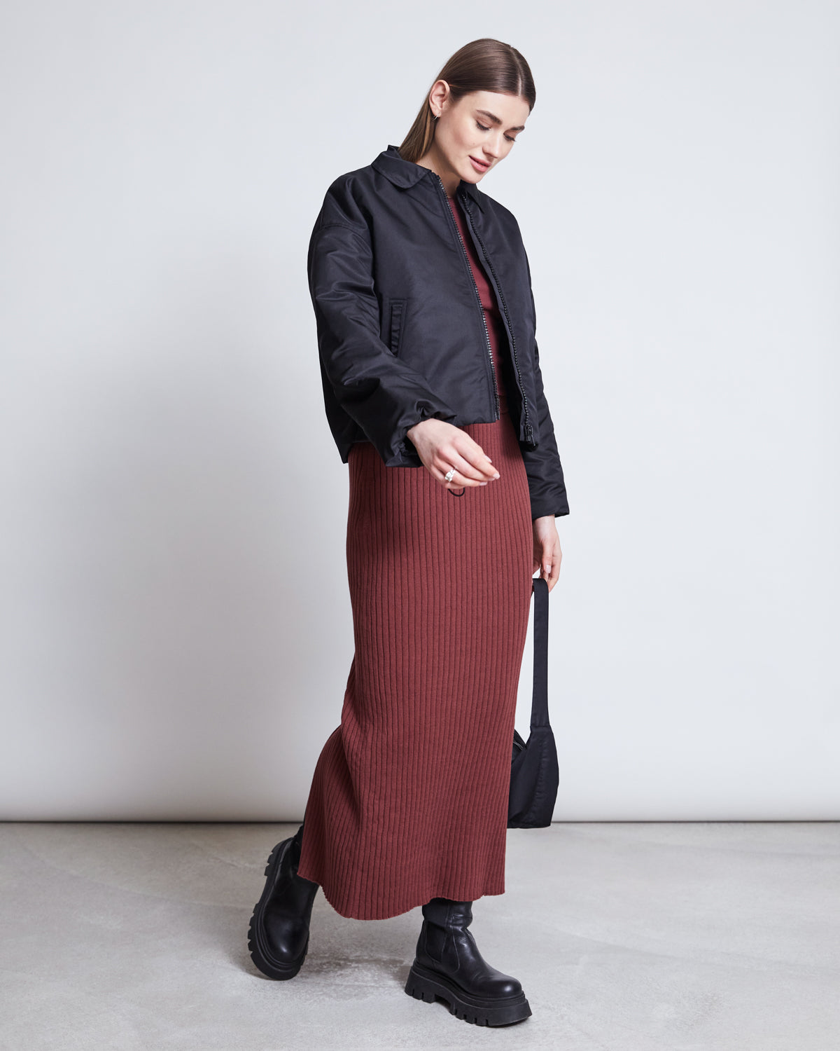 A woman stands against a plain background wearing the jan-n-june MIDI KNIT SKIRT EBBA INTENSE RUST GOTS, paired with a black bomber jacket, chunky black boots, and a small black bag. She looks down and smiles softly.