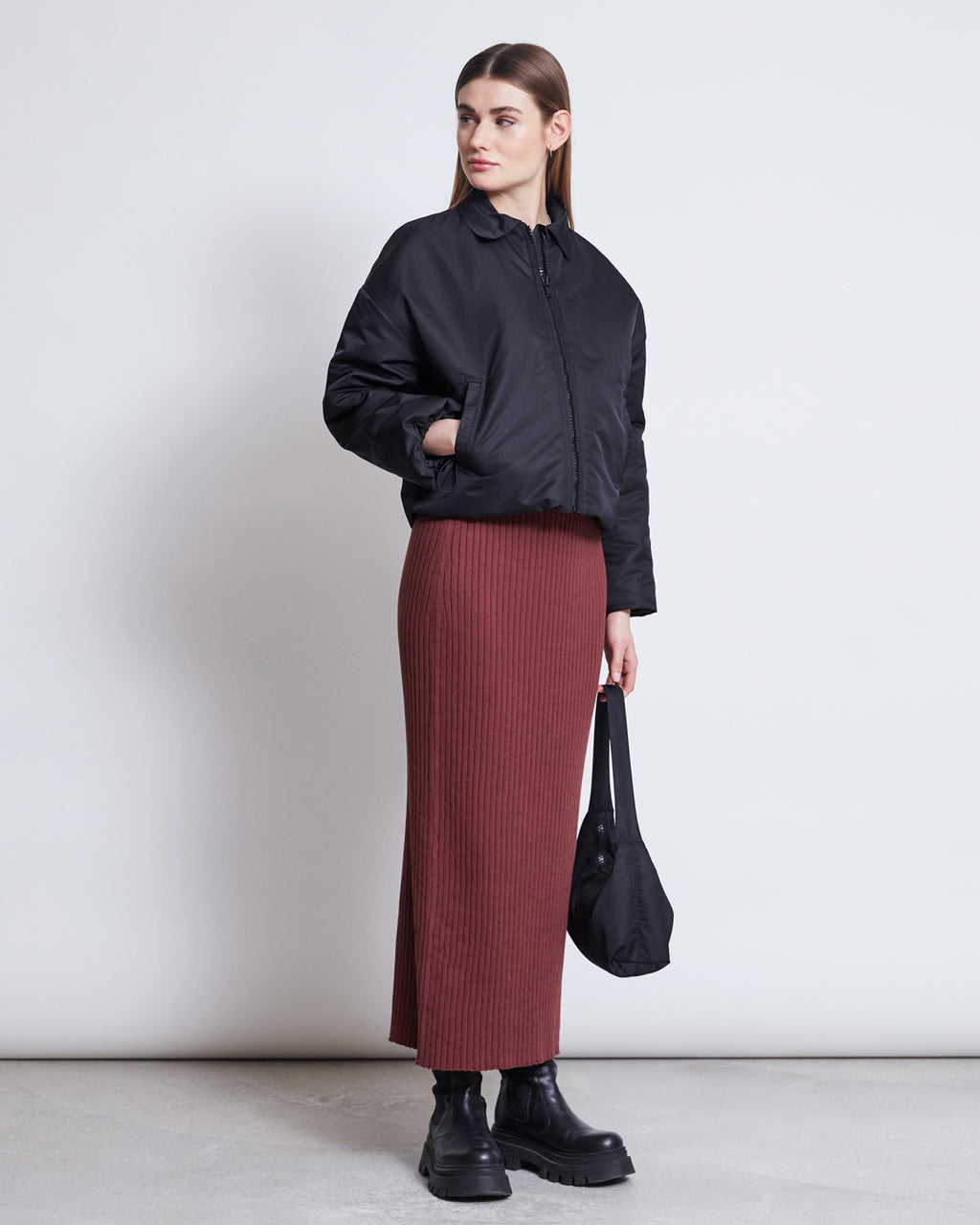 A woman stands against a plain background wearing the jan-n-june REC. BLOUSON DUSK BLACK jacket, a long maroon ribbed skirt, black chunky platform boots, and holding a black bag at her side.
