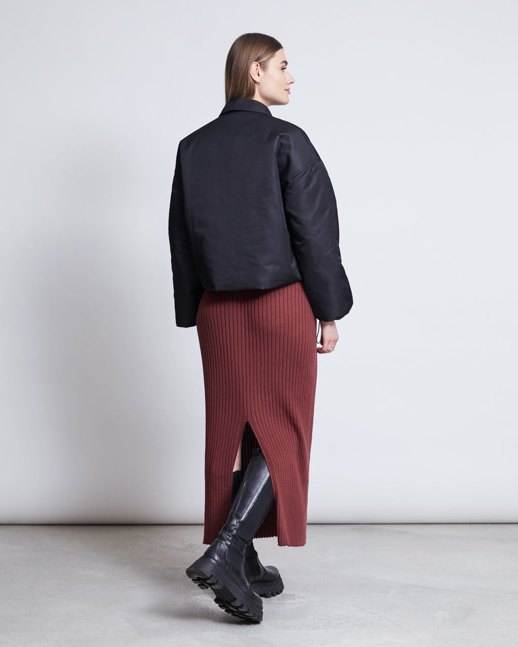 A woman with straight brown hair is standing with her back to the camera, wearing a black jacket, black knee-high boots, and the jan-n-june MIDI KNIT SKIRT EBBA INTENSE RUST GOTS, against a plain light background.