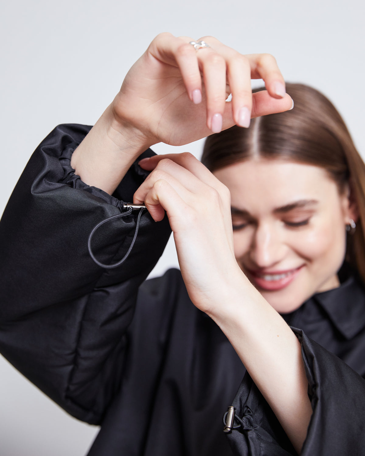 A woman with long brown hair smiles while adjusting the sleeve drawstring of the jan-n-june REC. BLOUSON DUSK BLACK, a sustainable jacket made from SEAQUAL® YARN, against a plain, light background. Her hands are near her face.