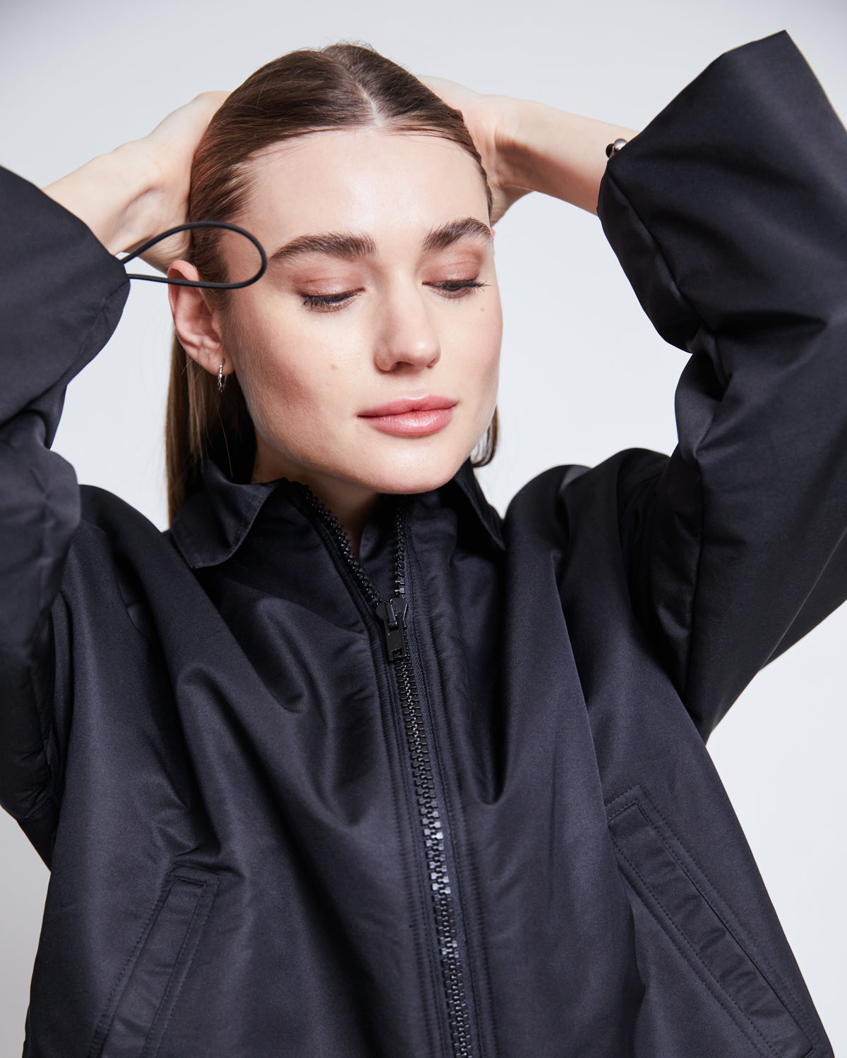 A woman with straight brown hair and closed eyes adjusts an elastic hair tie, wearing the jan-n-june REC. BLOUSON DUSK BLACK—a black zip-up jacket made from recycled material—while standing against a plain white background.