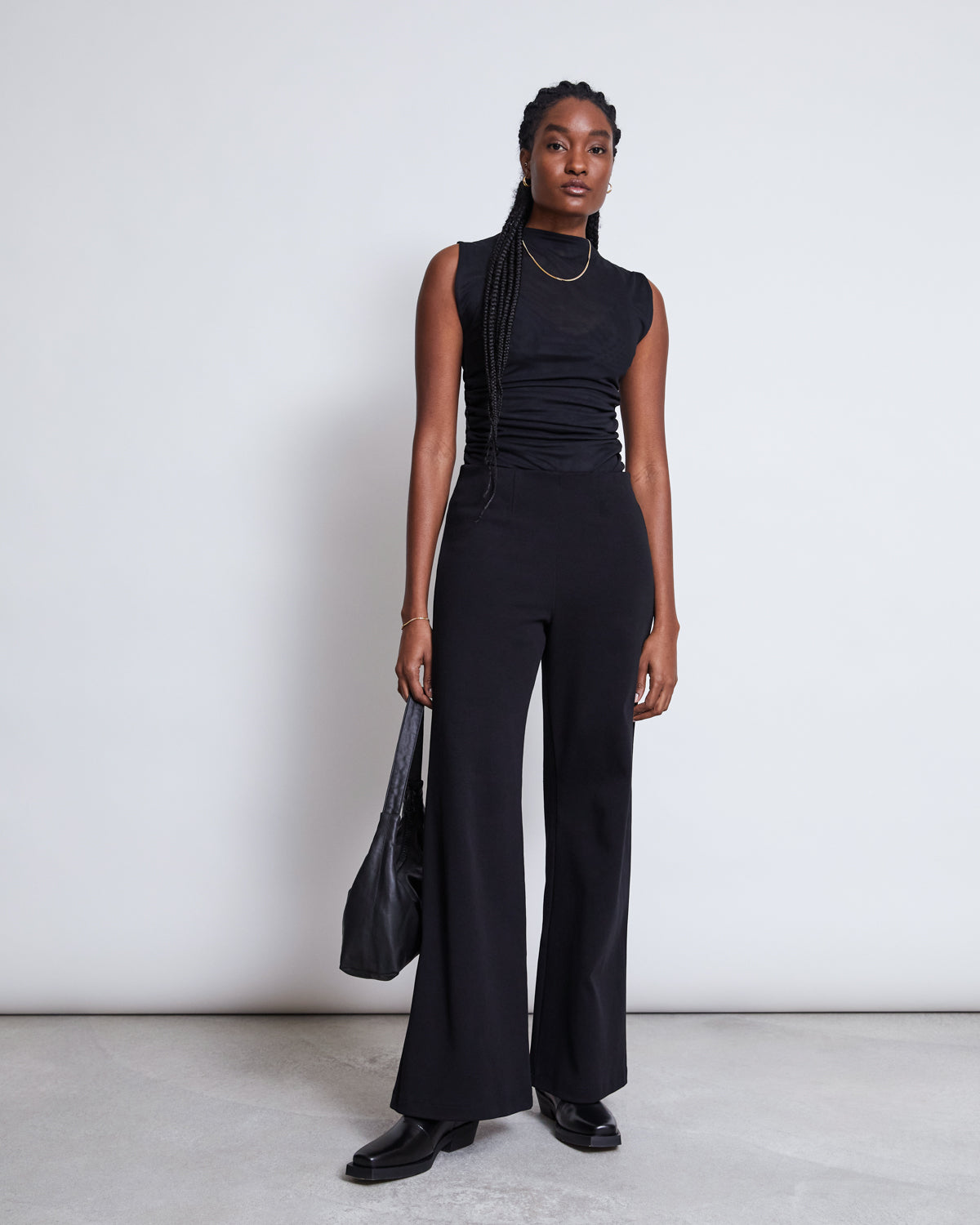 A woman with long braided hair and gold jewelry stands against a plain light background, wearing the jan-n-june RUFFLE MESH TOP LAGUNA BLACK, black flared pants, black shoes, and carrying a large black shoulder bag.