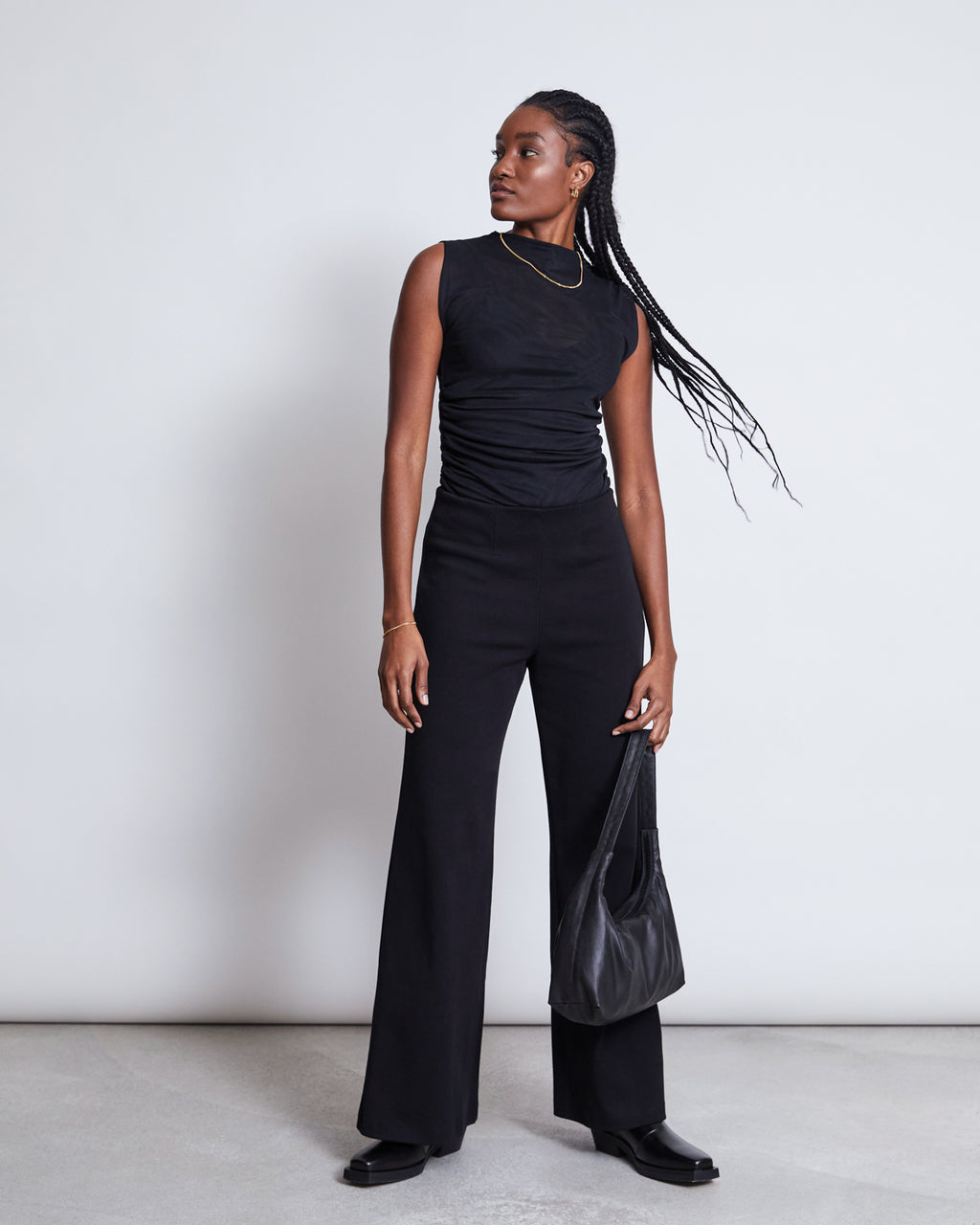 A woman with long braided hair over her shoulder stands against a plain light background, wearing the jan-n-june RUFFLE MESH TOP LAGUNA BLACK, black wide-leg pants, black shoes, and holding a black handbag while looking to her left.