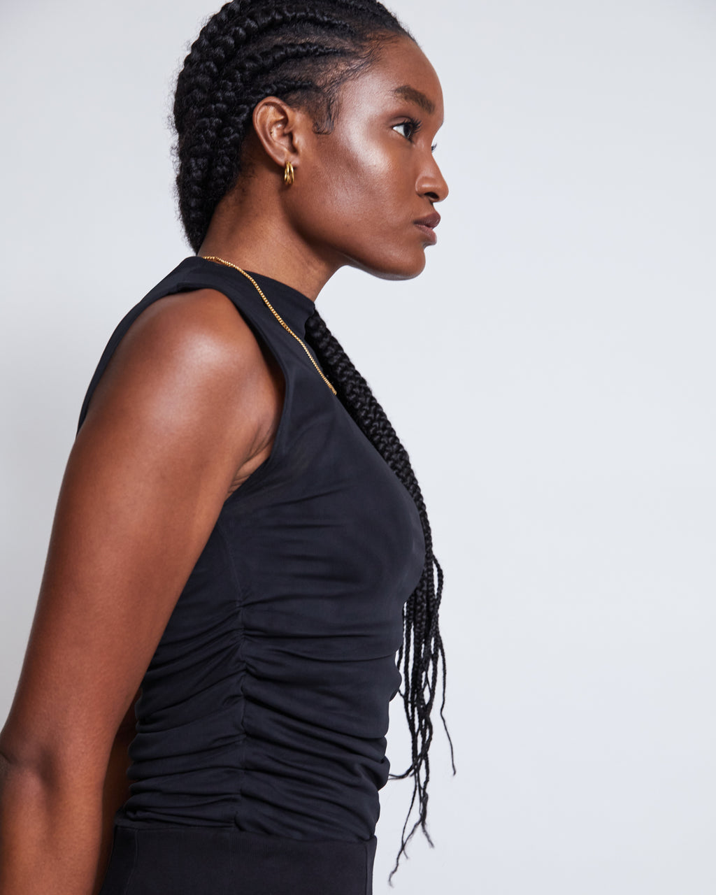 A woman with long braided hair, wearing the jan-n-june RUFFLE MESH TOP LAGUNA BLACK and gold hoop earrings, stands in profile against a plain white background.