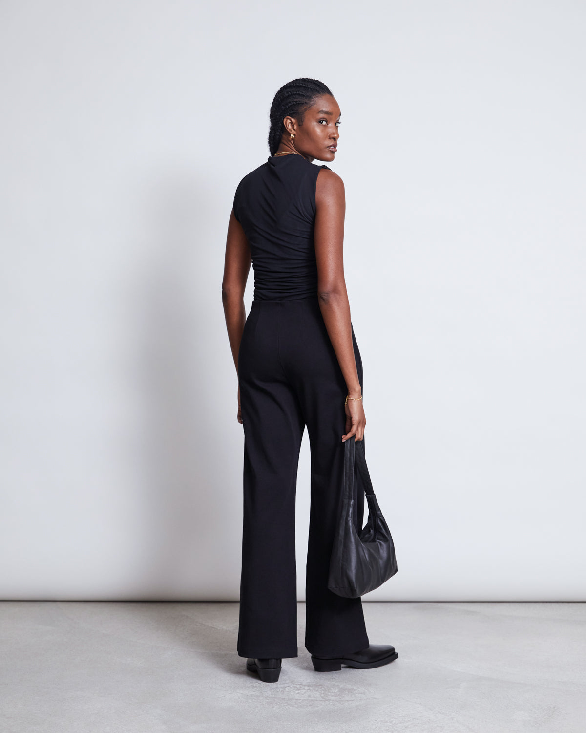 A woman with braided hair stands facing away, looking over her shoulder. She wears the jan-n-june RUFFLE MESH TOP LAGUNA BLACK, wide-leg black pants, black shoes, and holds a black bag against a plain light background.