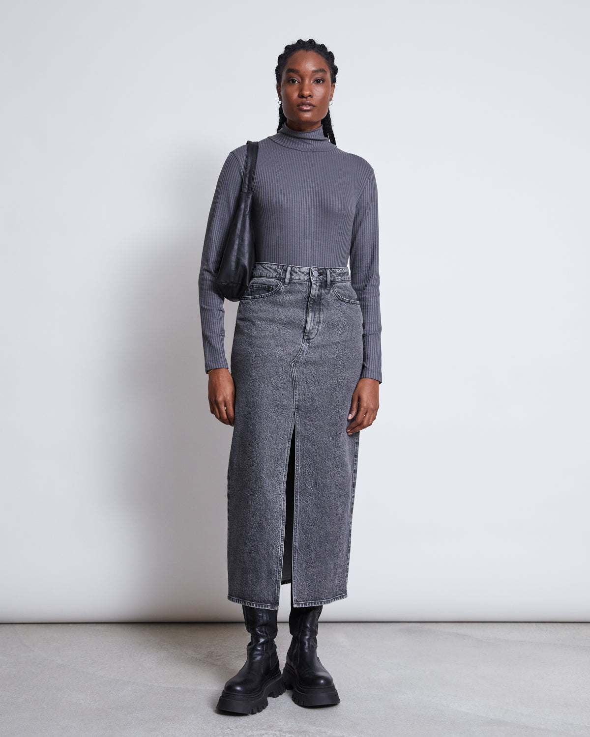A woman stands against a white background wearing the jan-n-june LYOCELL TURTLENECK MIO DARK GREY, paired with a high-waisted gray denim midi skirt with a front slit, black chunky boots, and a black shoulder bag.