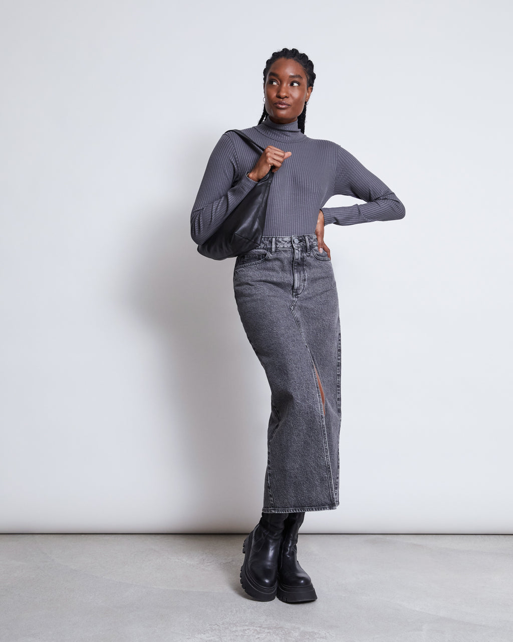 A woman stands against a plain white background, wearing the jan-n-june LYOCELL TURTLENECK MIO DARK GREY, a high-waisted gray denim midi skirt with a front slit, black boots, and holding a black shoulder bag.