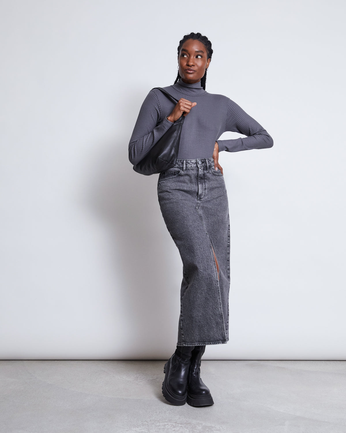 A woman stands against a plain white background, wearing the jan-n-june LYOCELL TURTLENECK MIO DARK GREY, a high-waisted gray denim midi skirt with a front slit, black boots, and holding a black shoulder bag.
