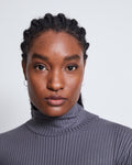 A woman with braided hair and small hoop earrings wears the jan-n-june LYOCELL TURTLENECK MIO DARK GREY, looking directly at the camera with a neutral expression against a plain light background.