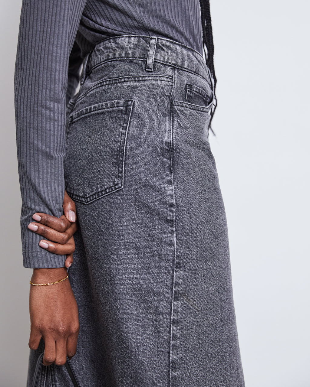 A person stands sideways, hand on hip, wearing jan-n-junes LYOCELL TURTLENECK MIO DARK GREY paired with a high-waisted, faded gray denim skirt featuring visible stitching and belt loops.