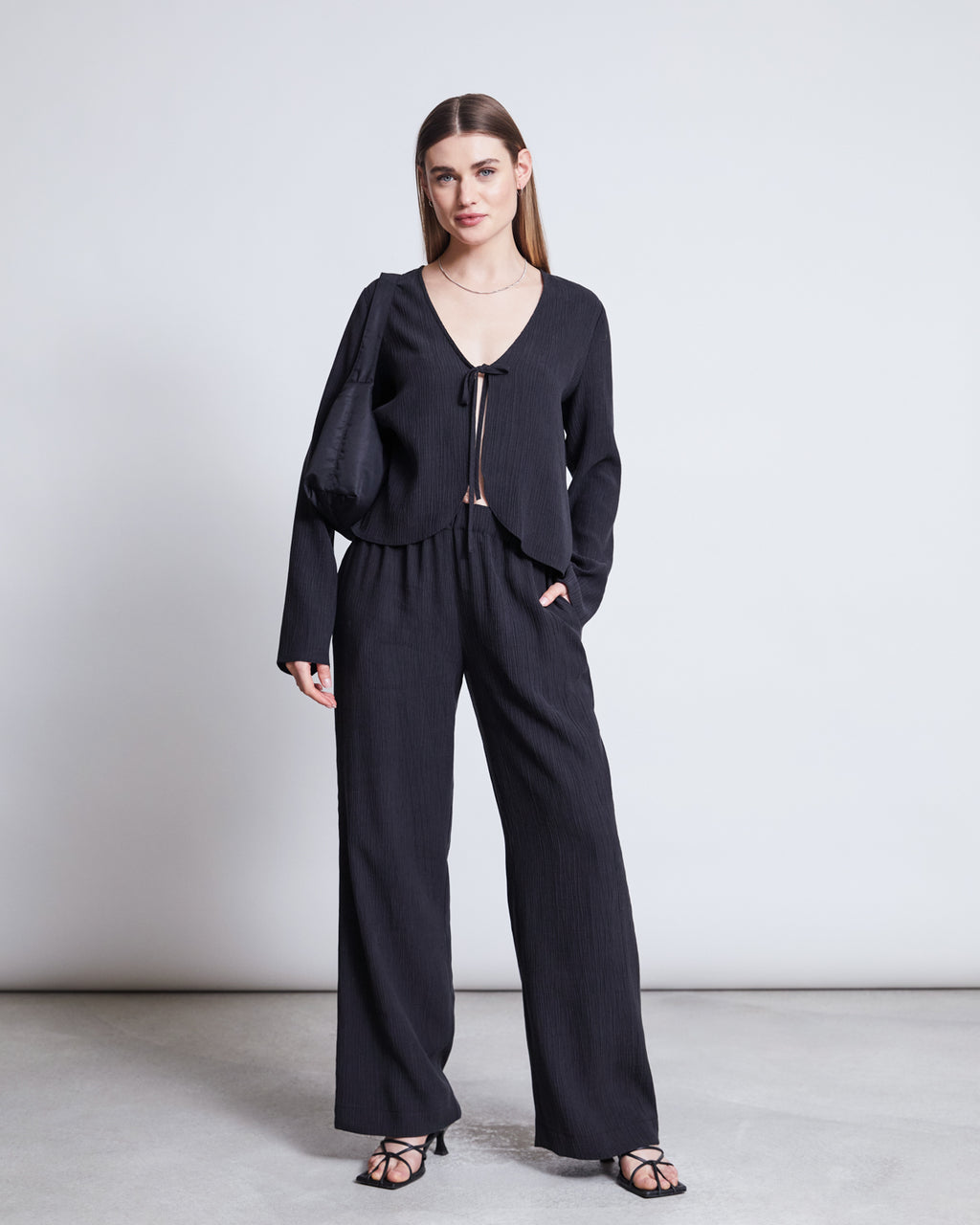 A woman with straight hair stands against a plain background wearing jan-n-june’s CRINKLE PANTS HAVA BLACK and a loose black top with front tie, paired with sandals and a black shoulder bag.