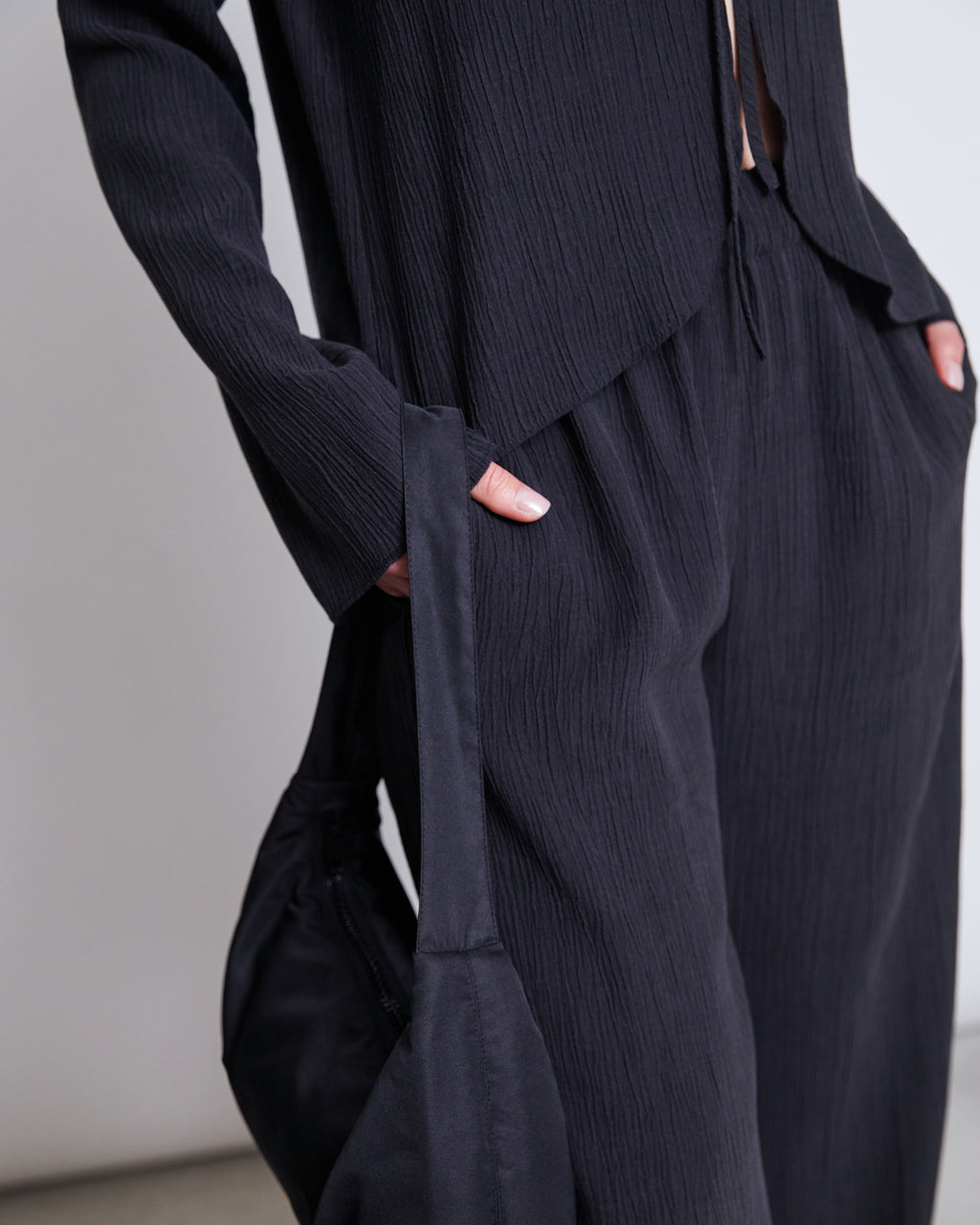 A person wears jan-n-junes CRINKLE PANTS HAVA BLACK matching set—wide-leg crinkle pants and a long-sleeve top. With hands in their pockets, they carry a black bag over one arm. The image is cropped at the shoulders.