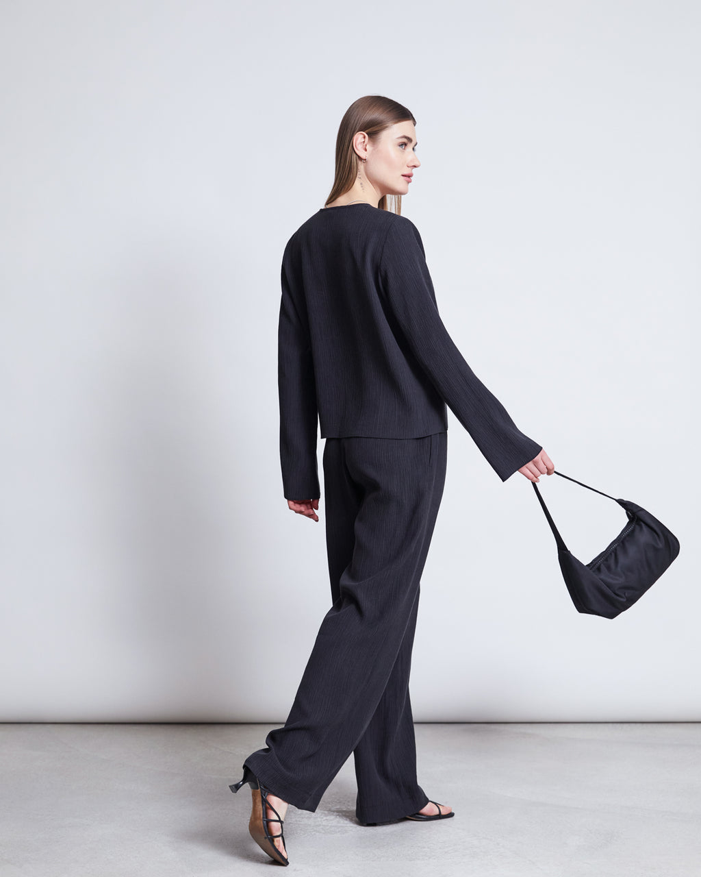 A woman with straight brown hair models jan-n-junes CRINKLE PANTS HAVA BLACK with sandals and a small black handbag, glancing over her shoulder as she walks away against a white background.