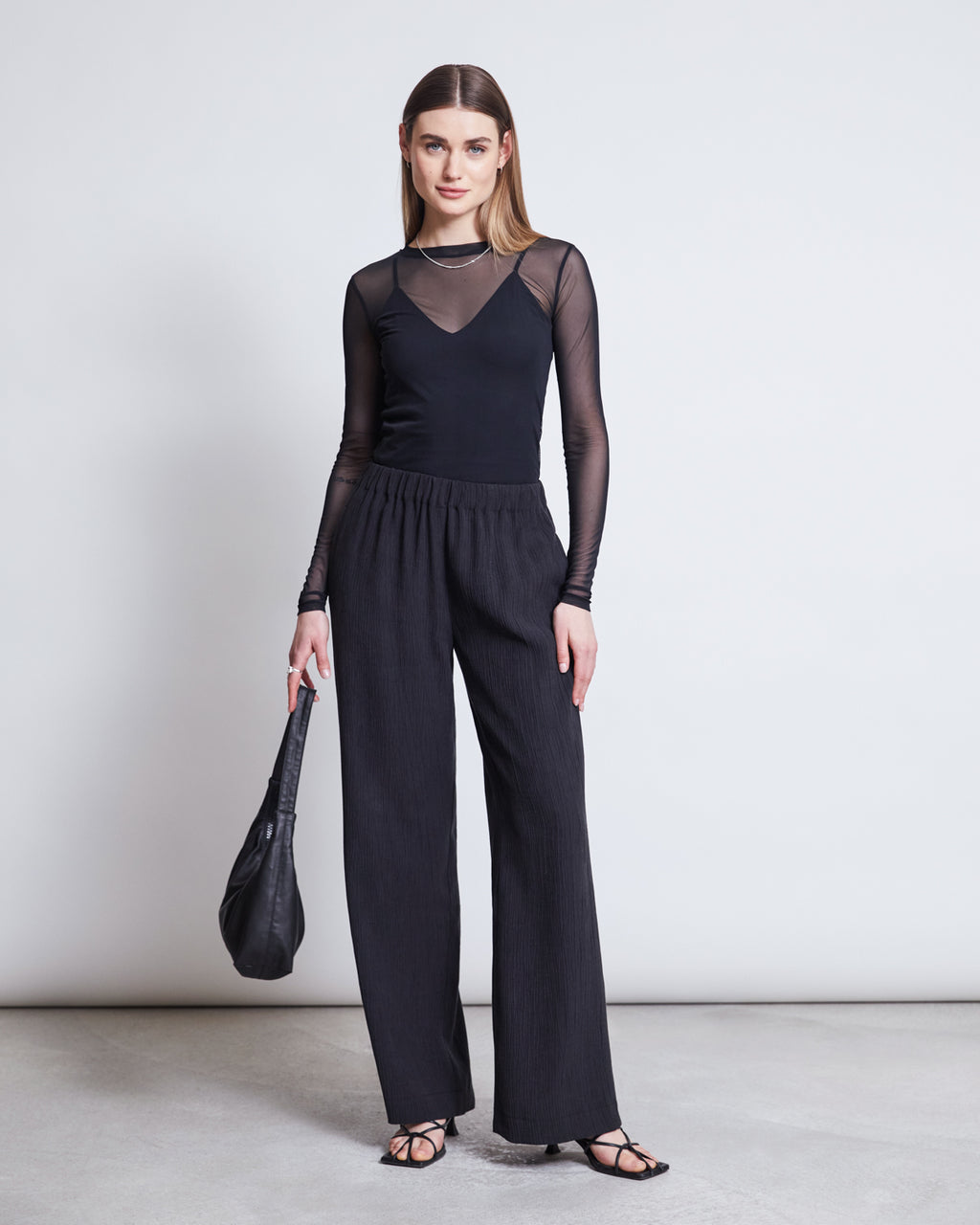 A woman with straight, shoulder-length hair and a neutral expression stands against a plain background wearing the jan-n-june MESH LONGSLEEVE DORA BLACK top, black wide-leg pants, black sandals, and holding a black bag.