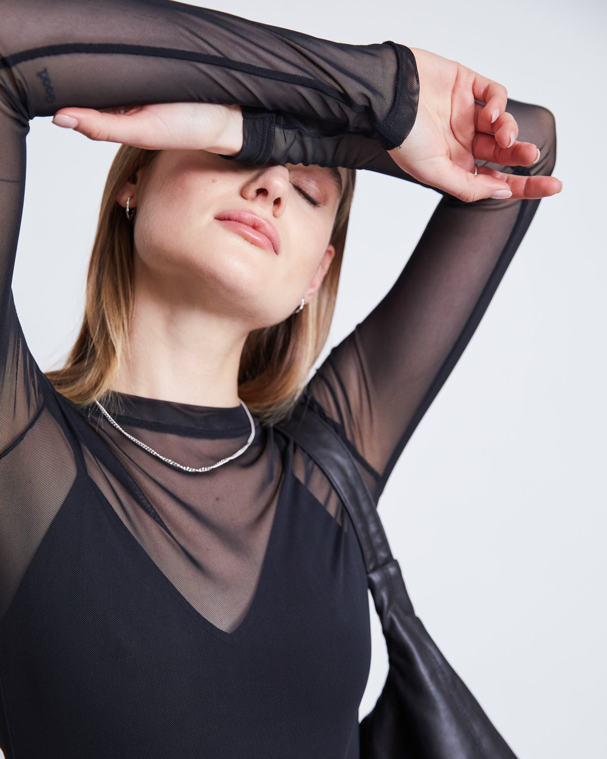 Wearing the jan-n-june MESH LONGSLEEVE DORA BLACK, made from recycled plastic bottles, and a silver necklace, a person poses with one arm across their forehead, eyes closed, holding a black bag over their shoulder.