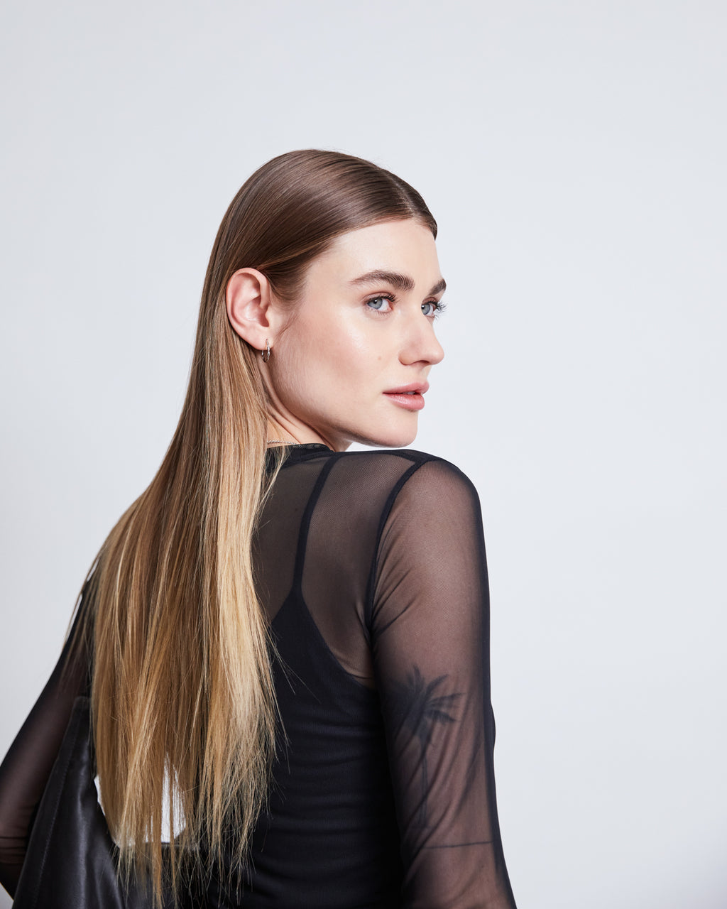 A person with long, straight, light brown hair looks over their shoulder, wearing the jan-n-june MESH LONGSLEEVE DORA BLACK made from recycled plastic bottles, set against a plain light background.