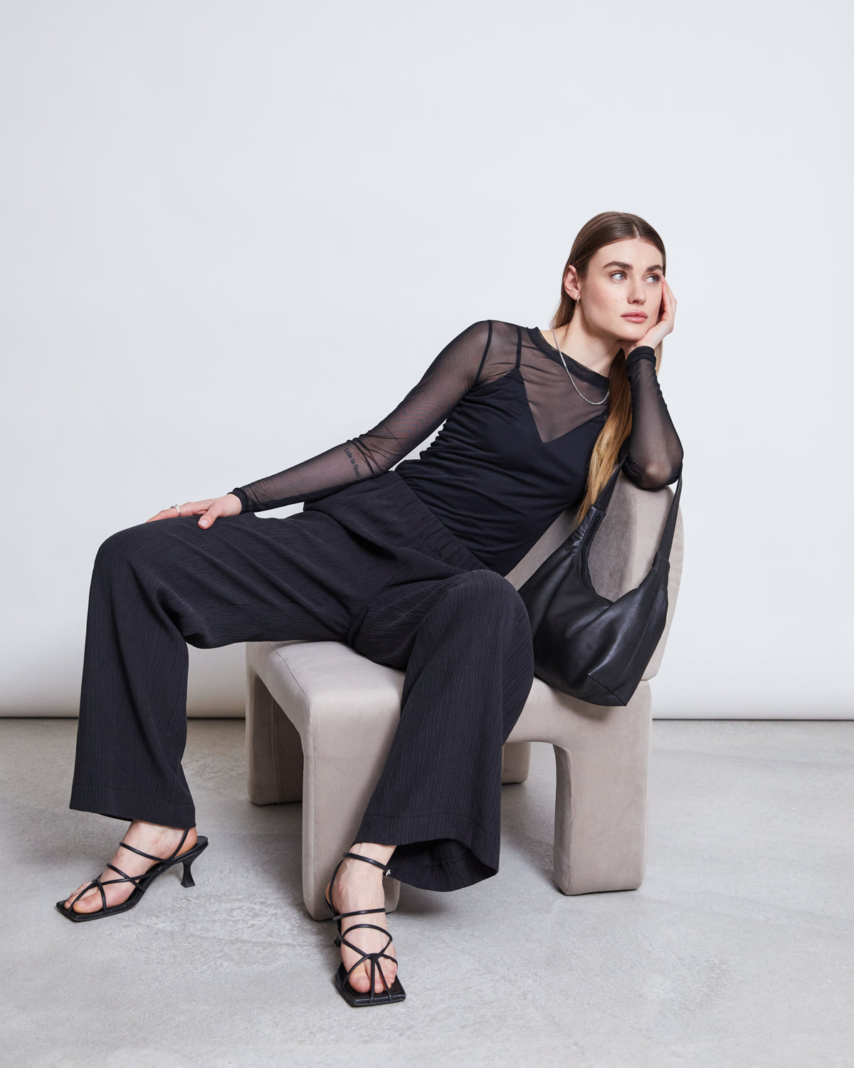 A woman wearing jan-n-june CRINKLE PANTS HAVA BLACK, a sheer black top, and strappy heels sits on a modern chair with a relaxed expression, straight hair, and a black handbag on her arm.