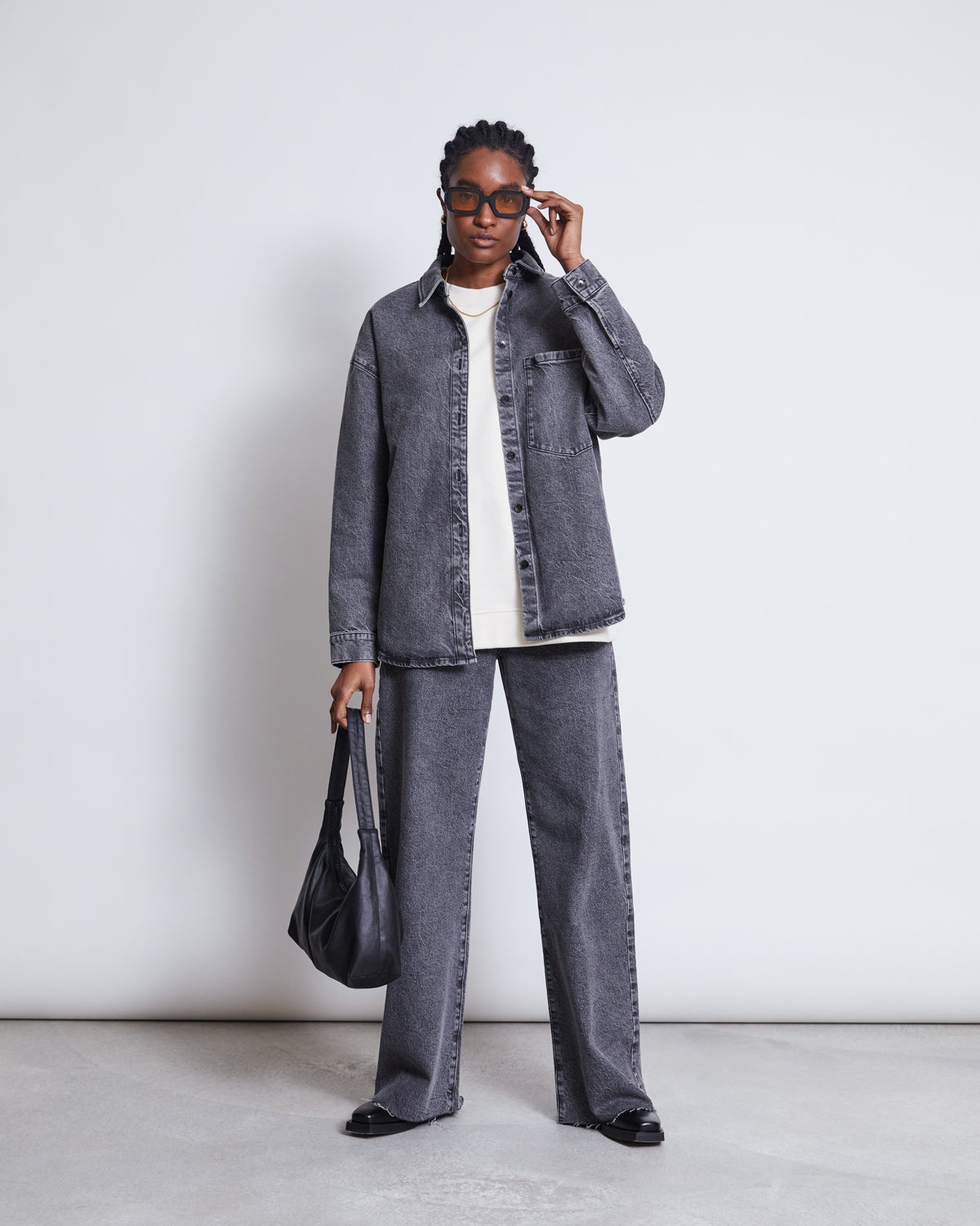 A person wearing the jan-n-june DENIM SHIRT SOLACE LIGHT GREY, matching wide-leg pants, a white shirt, black shoes, sunglasses, and holding a black handbag stands against a plain white background.