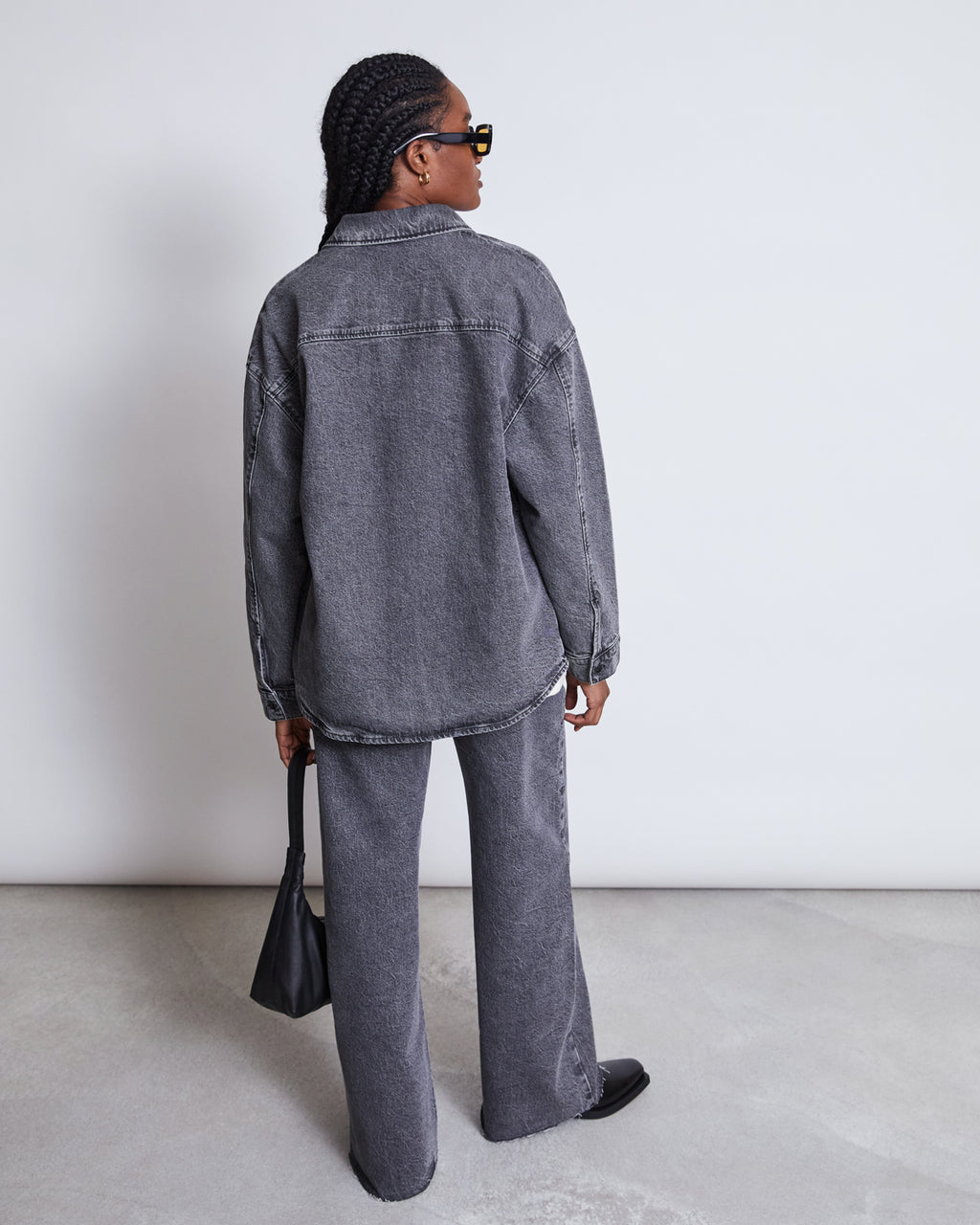 A person with braided hair faces away from the camera, wearing jan-n-junes DENIM SHIRT SOLACE LIGHT GREY in an oversized fit with matching pants, paired with a black handbag and shoes against a plain light background.