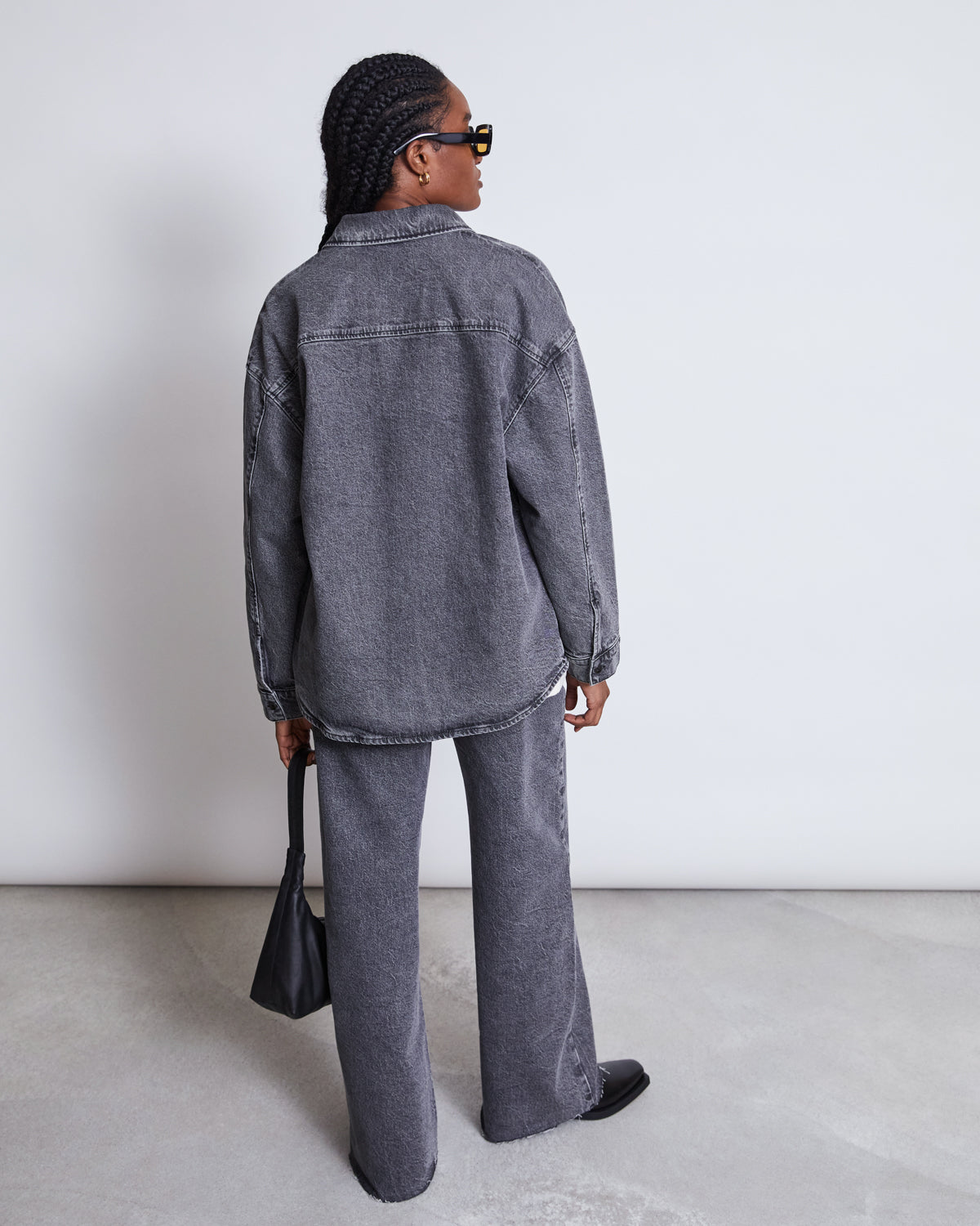 A person with braided hair faces away from the camera, wearing jan-n-junes DENIM SHIRT SOLACE LIGHT GREY in an oversized fit with matching pants, paired with a black handbag and shoes against a plain light background.