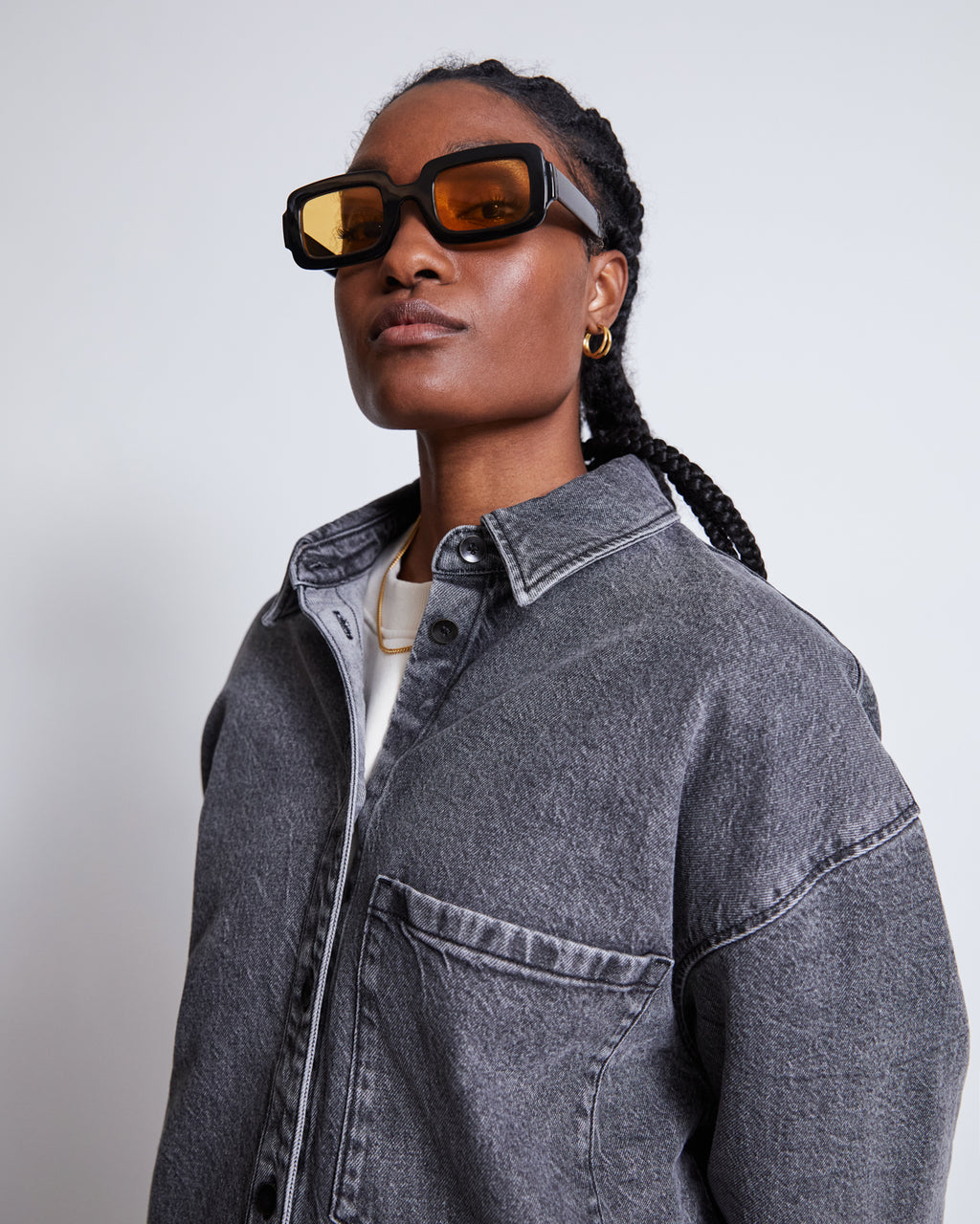 A person with braided hair wears chunky brown sunglasses, gold hoop earrings, and the jan-n-june DENIM SHIRT SOLACE LIGHT GREY over a white tee, standing against a plain light background.