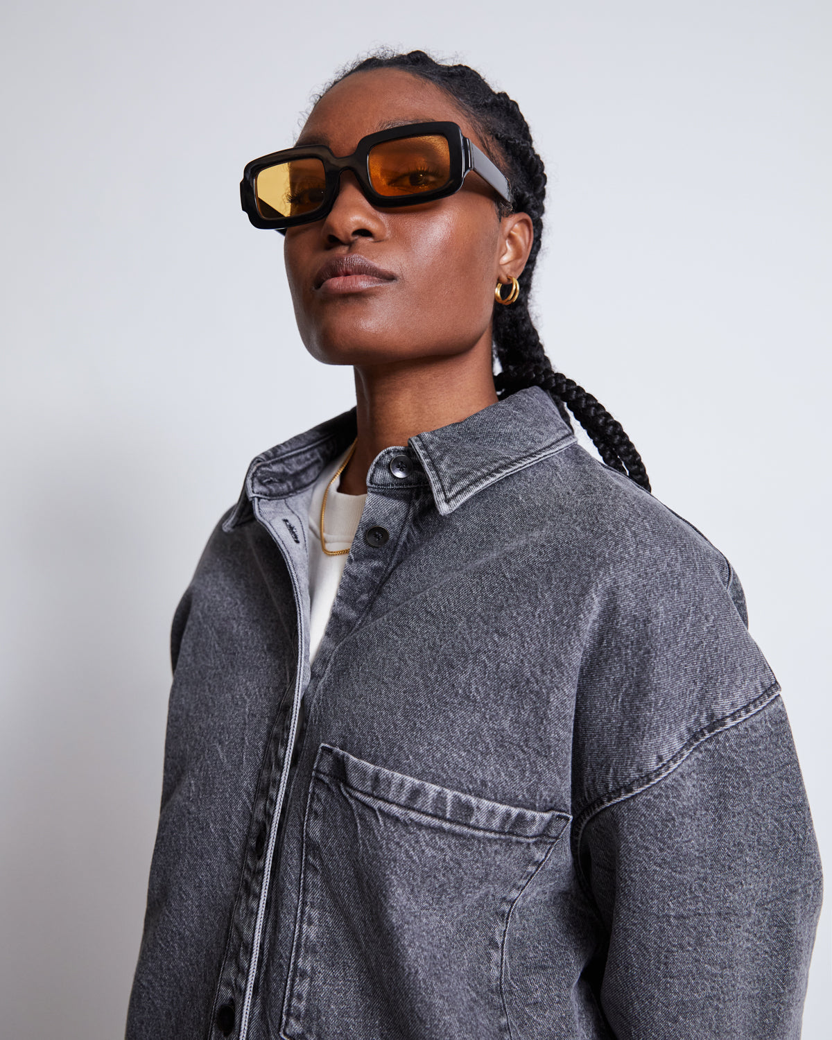 A person with braided hair wears chunky brown sunglasses, gold hoop earrings, and the jan-n-june DENIM SHIRT SOLACE LIGHT GREY over a white tee, standing against a plain light background.