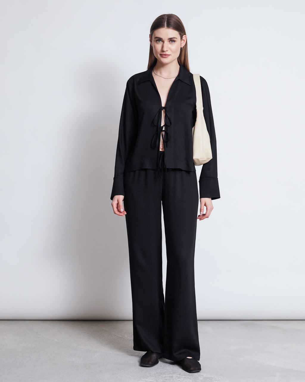 A woman with long straight hair and a neutral expression stands against a white wall, wearing the jan-n-june TIE BLOUSE KALUZ BLACK with matching wide-leg pants, black shoes, and carrying a light beige shoulder bag.