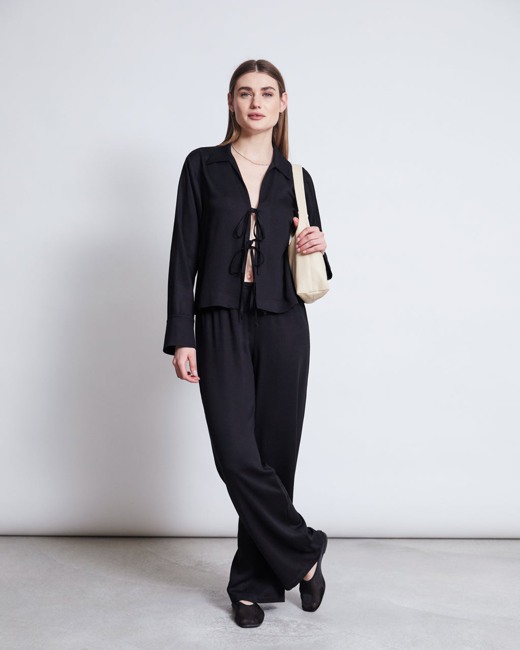 A woman stands against a plain white background wearing the jan-n-june TIE BLOUSE KALUZ BLACK and matching wide-leg pants. She carries a light beige shoulder bag, wears black shoes, and smiles softly with her left hand by her side.