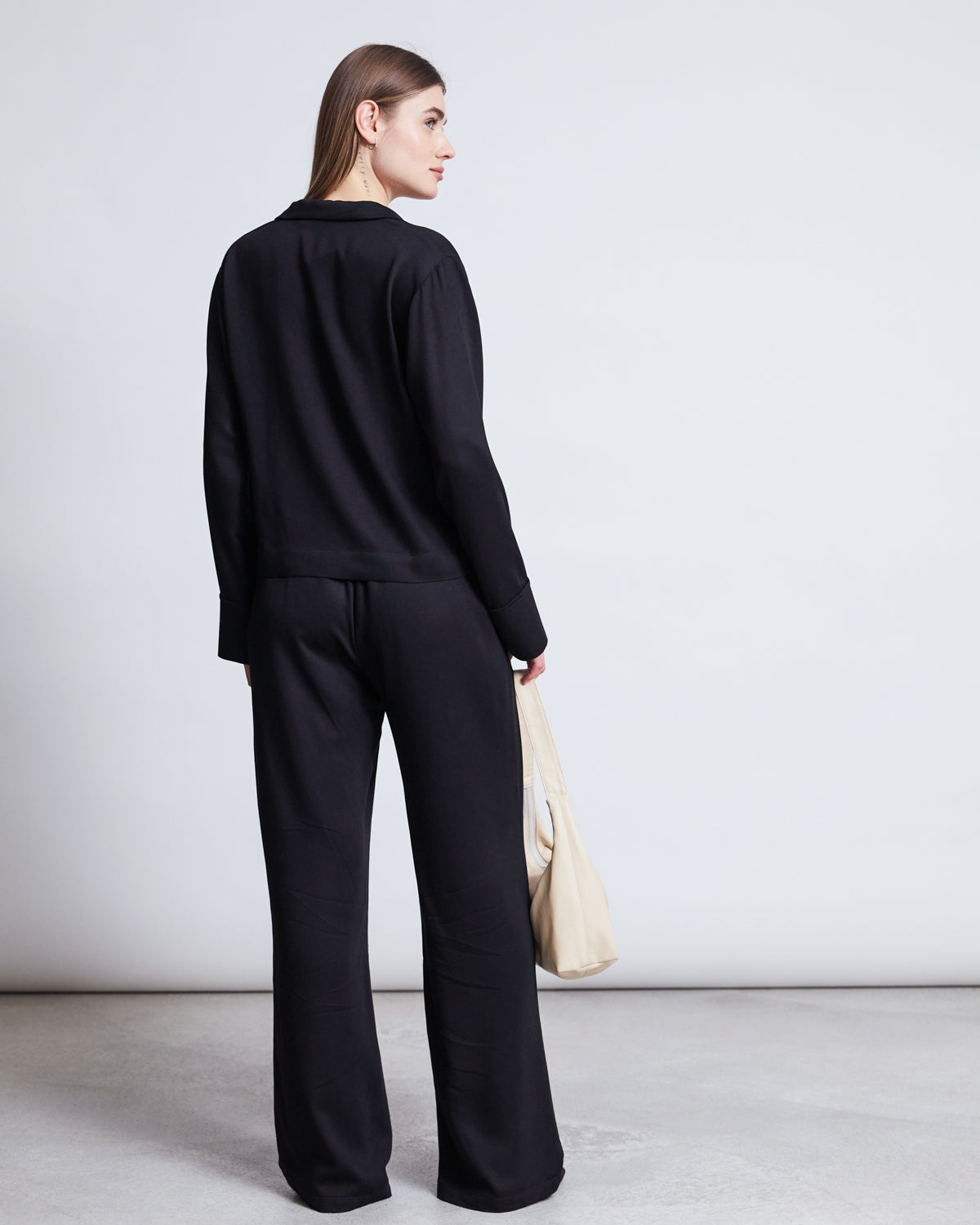 A woman faces away, wearing the jan-n-june TIE BLOUSE KALUZ BLACK and matching black pants. She holds a light-colored bag in her left hand, standing against a plain, light background.