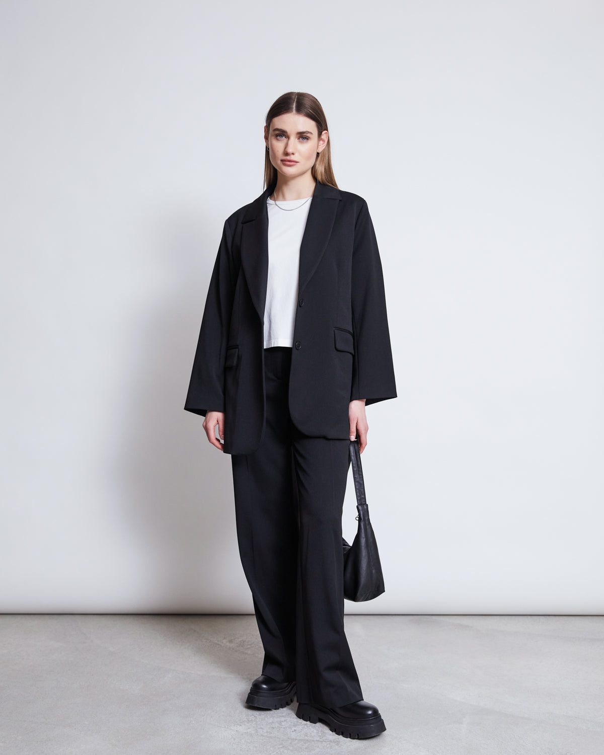 A woman with straight hair and a neutral expression stands against a white background wearing jan-n-june’s BLAZER DURANCE BLACK, matching wide-leg pants, a white t-shirt, black chunky shoes, and holding a black handbag.